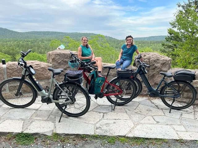 Zwei Frauen sitzen auf Felsen neben ihren geparkten E-Bikes und blicken über ein Tal.